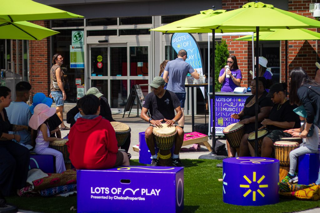 Community members gather in a circle playing hand drums on a grassy plaza during an outdoor Lots of Play event, with seating and activity stations nearby.