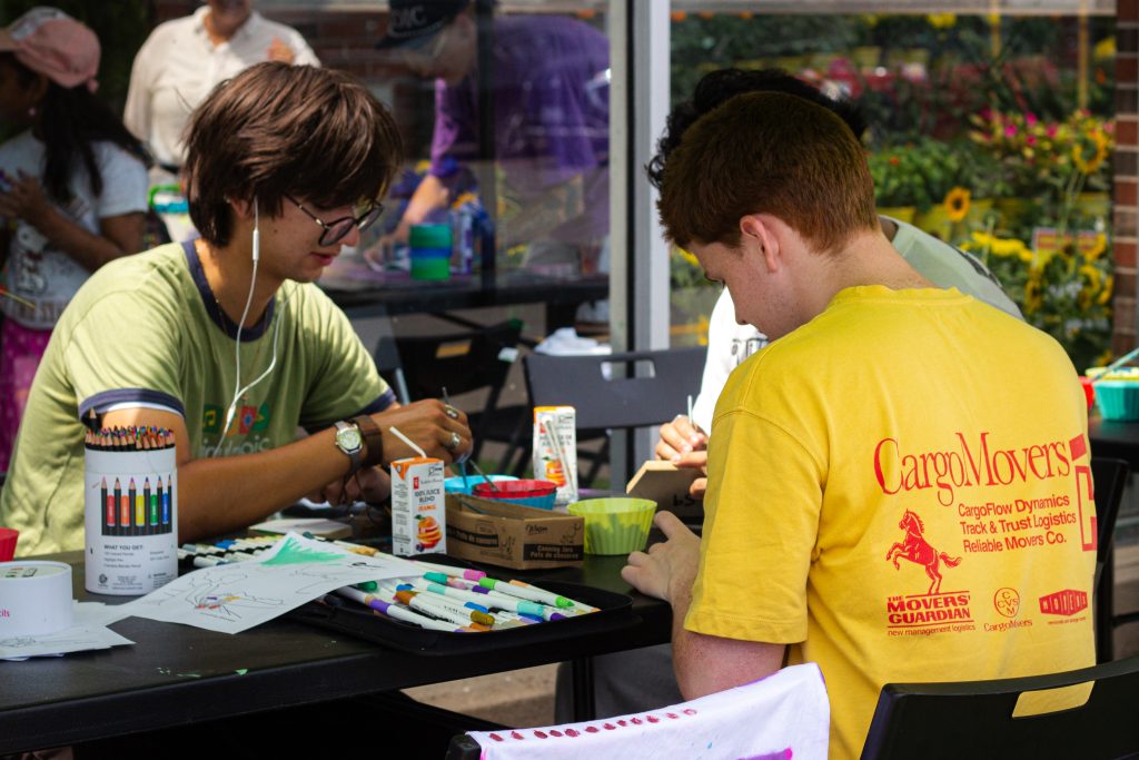Participants sit around a table drawing and colouring artwork with markers and pencils during an outdoor arts-and-crafts activity.