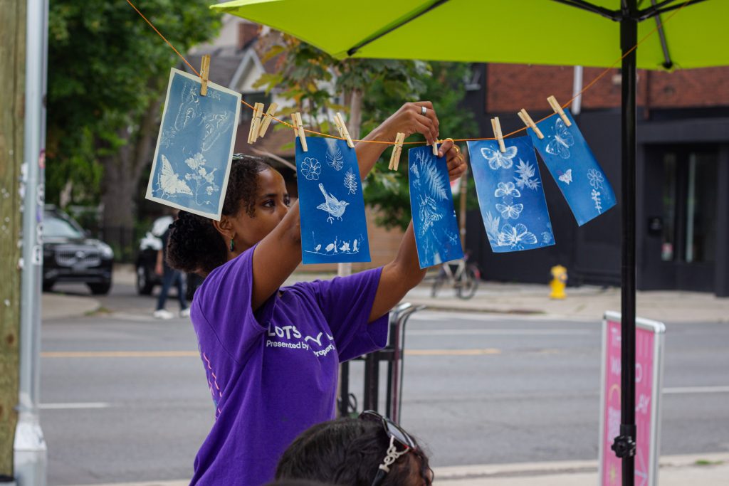 A participant hangs blue cyanotype prints featuring botanical and nature-inspired designs on a clothesline display beneath a bright green umbrella at an outdoor community art event.